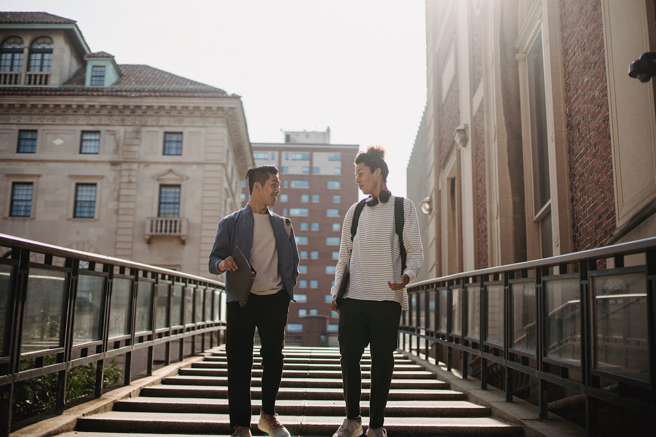 Two young men walking and talking on campus stairs under sunlight.